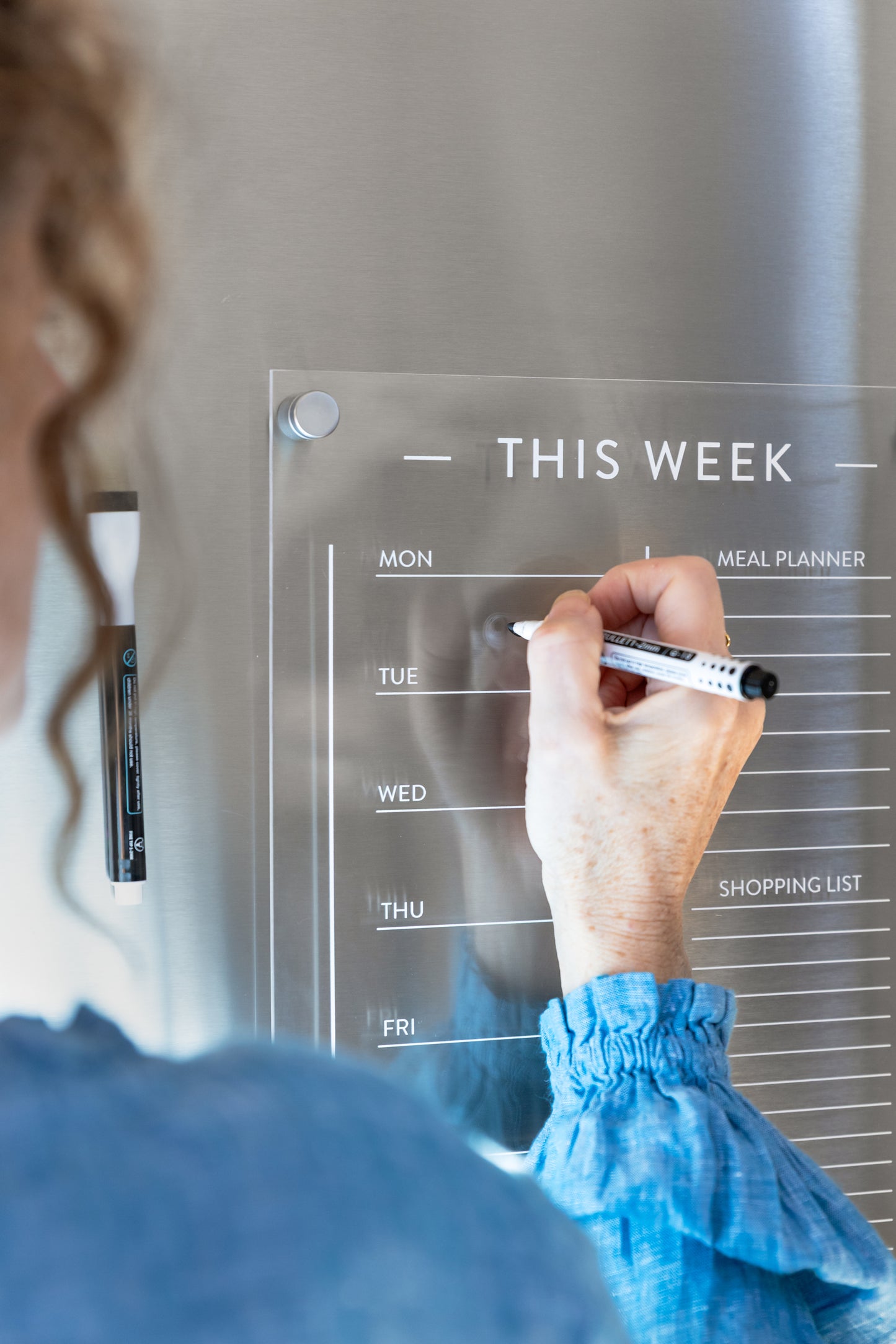 Person writing on a luxury acrylic magnetic meal planner mounted on a fridge, styled in a New Zealand kitchen.