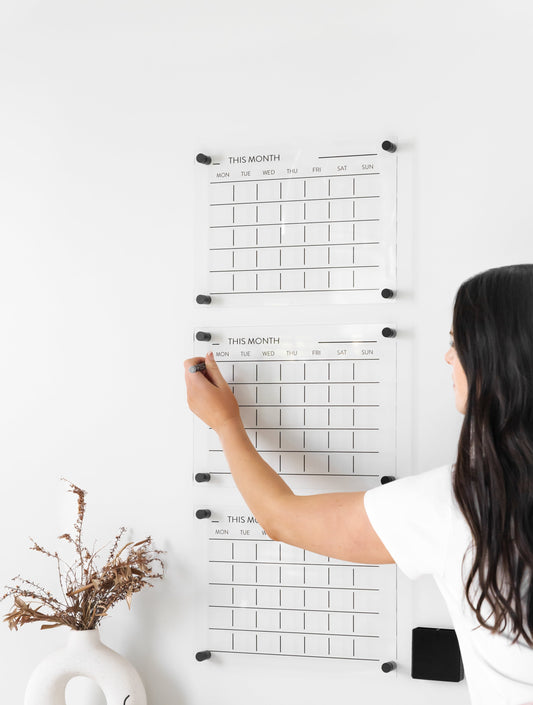 Person writing on a triple month acrylic wall planner mounted on a white wall, ideal for family scheduling and shift work organisation